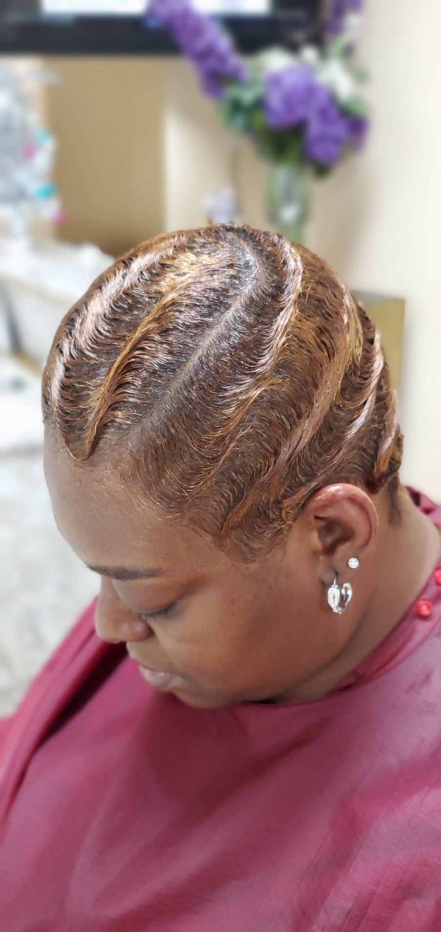 Side view of braided hair with polished swirls, shown against a blurred floral salon background