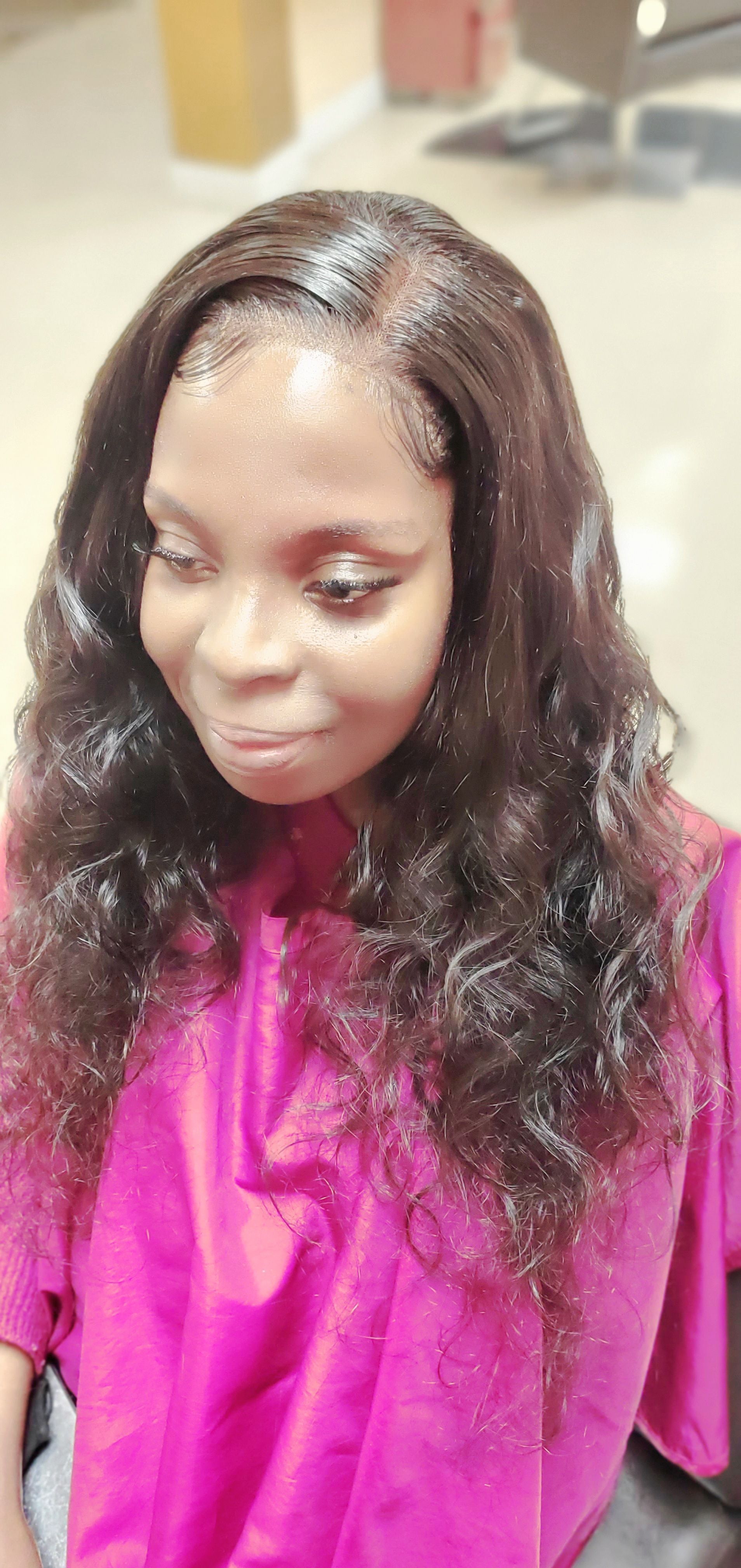 Person with long wavy hair wearing a bright pink top, looking down in a salon chair.