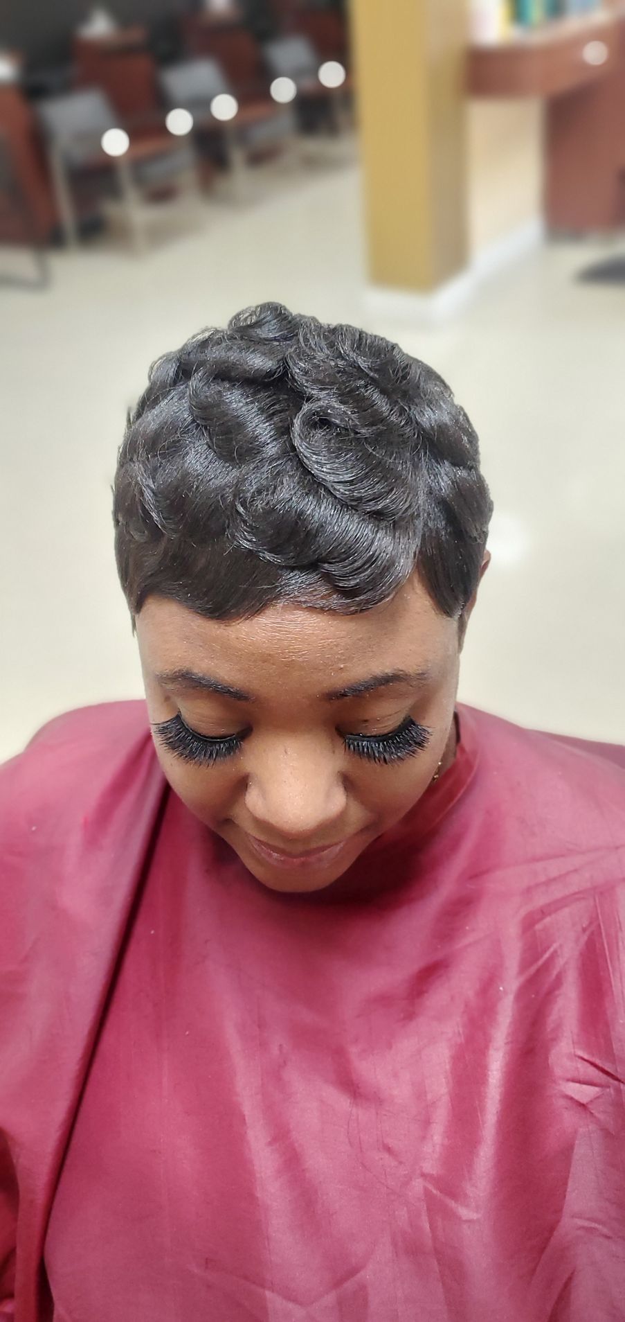 Person with short dark curls looking down, wearing a maroon salon cape in a bright salon.