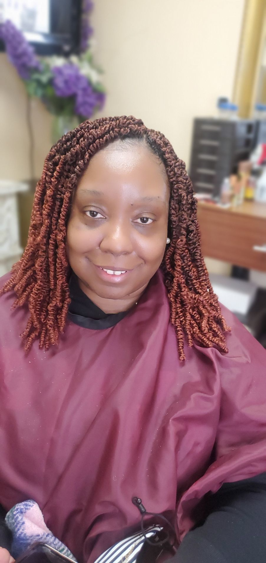 Person seated in a salon chair, smiling, wearing a burgundy cape and braided hair.
