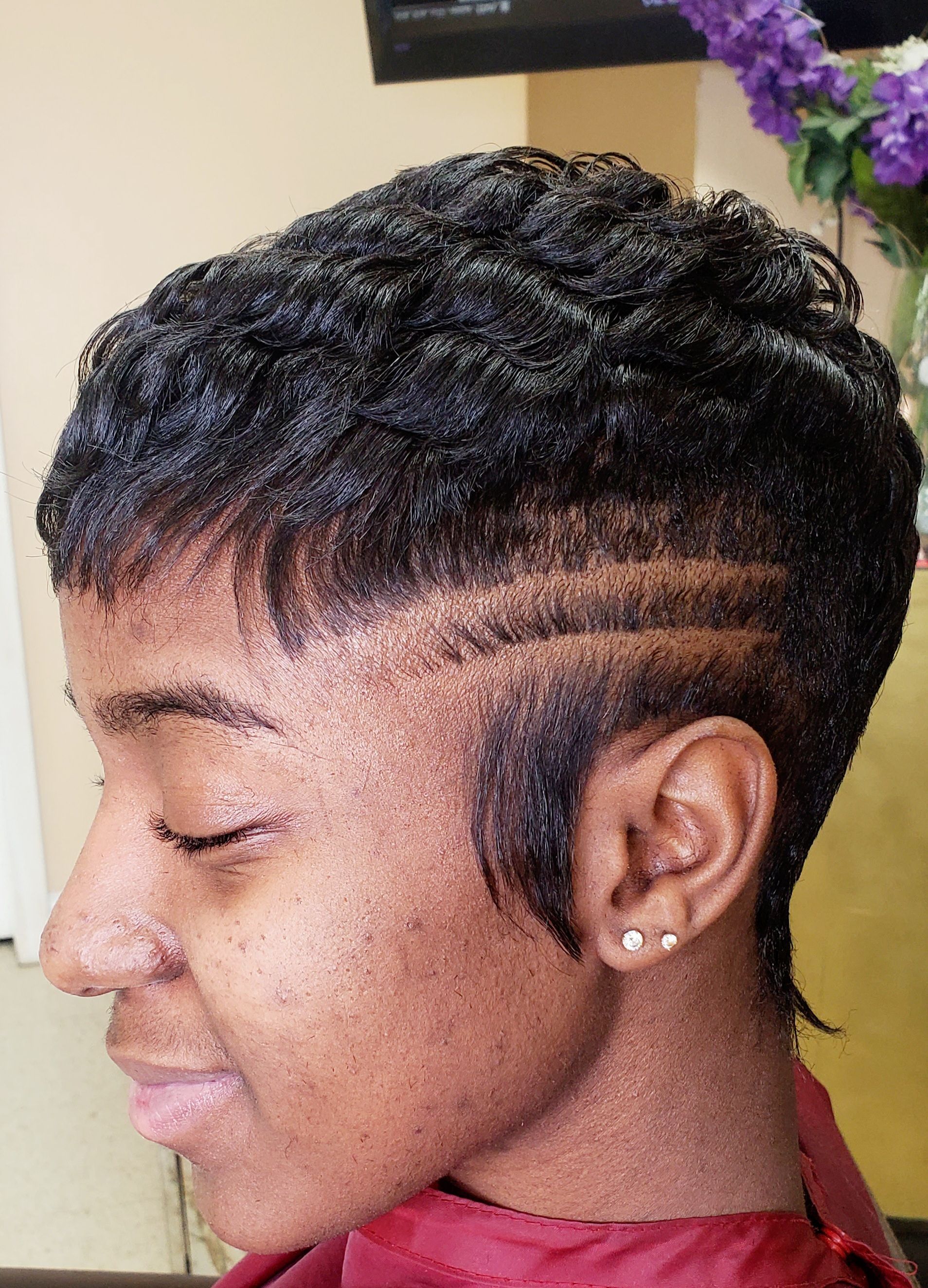 Profile view of a person with short braided hair and shaved side, wearing an earring, smiling slightly