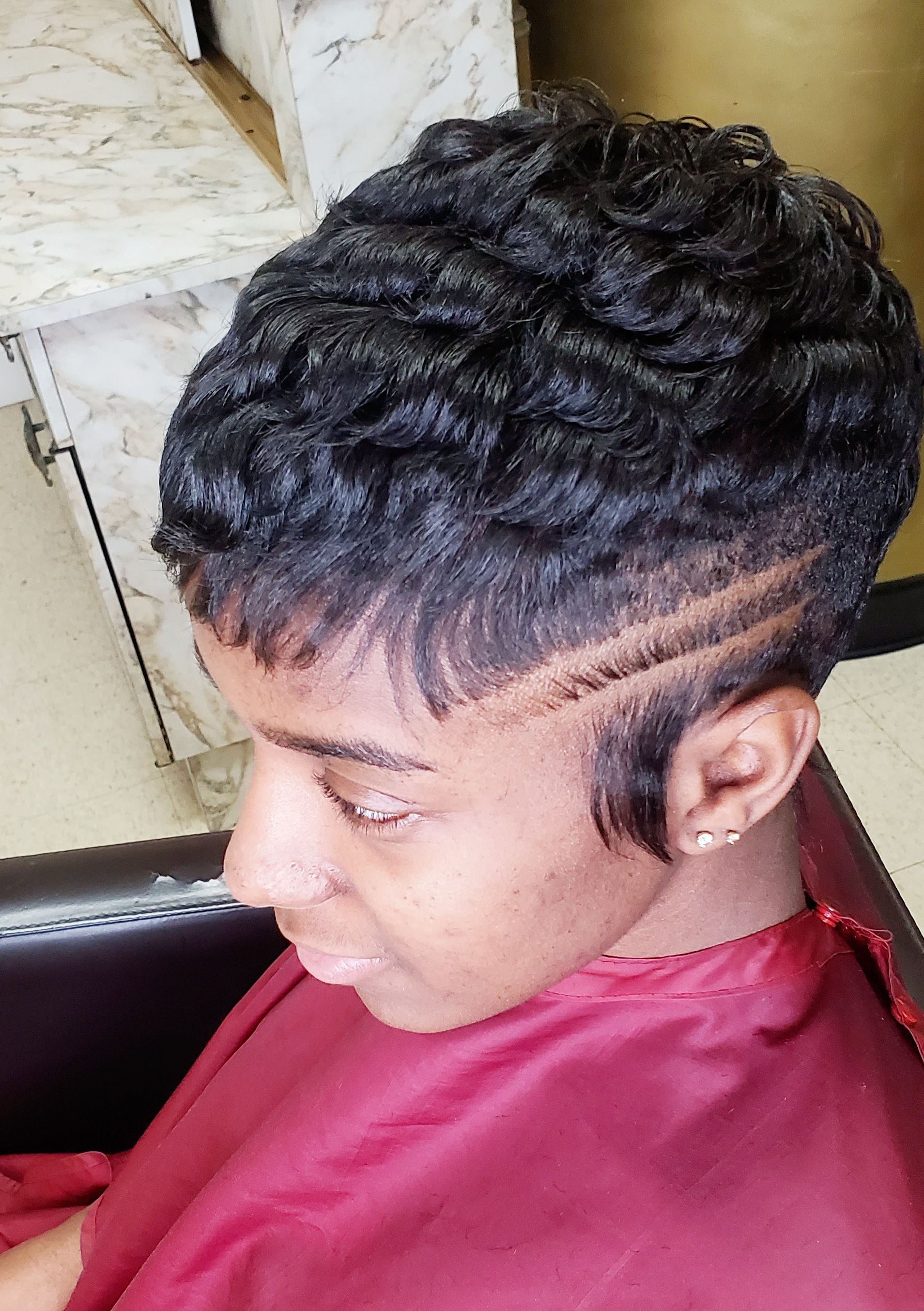 Short curly hairstyle with side designs on a person in a maroon barber cape