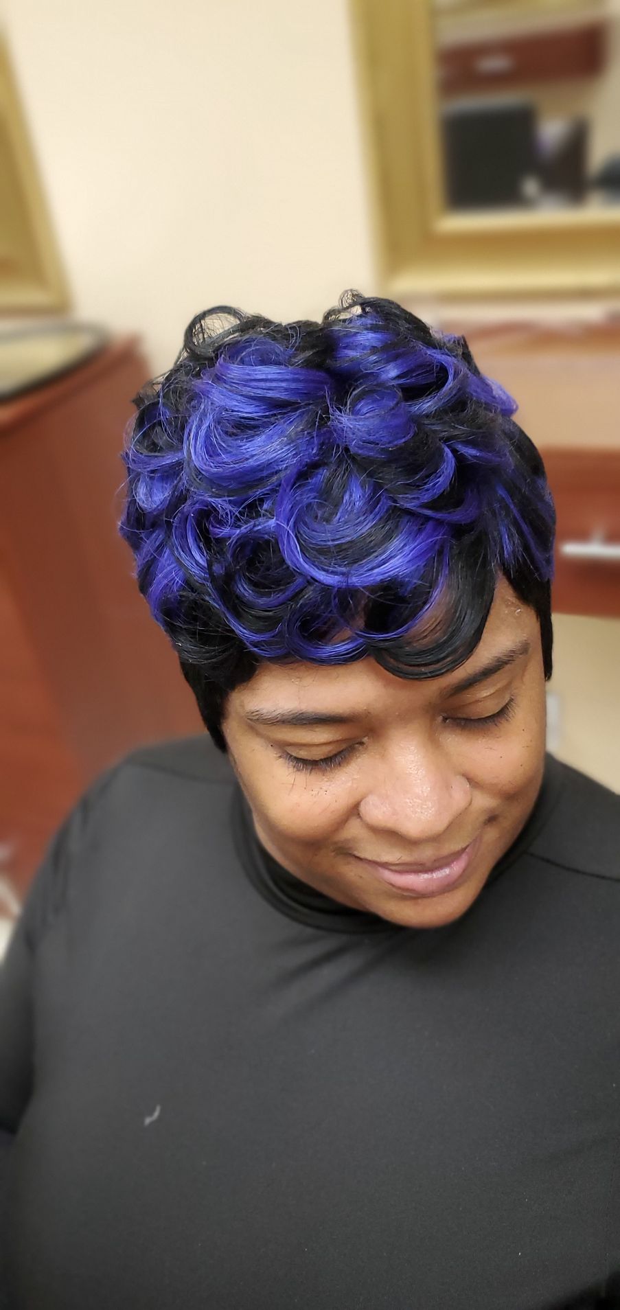 Person with short curly purple hair looking down, wearing a black top indoors.
