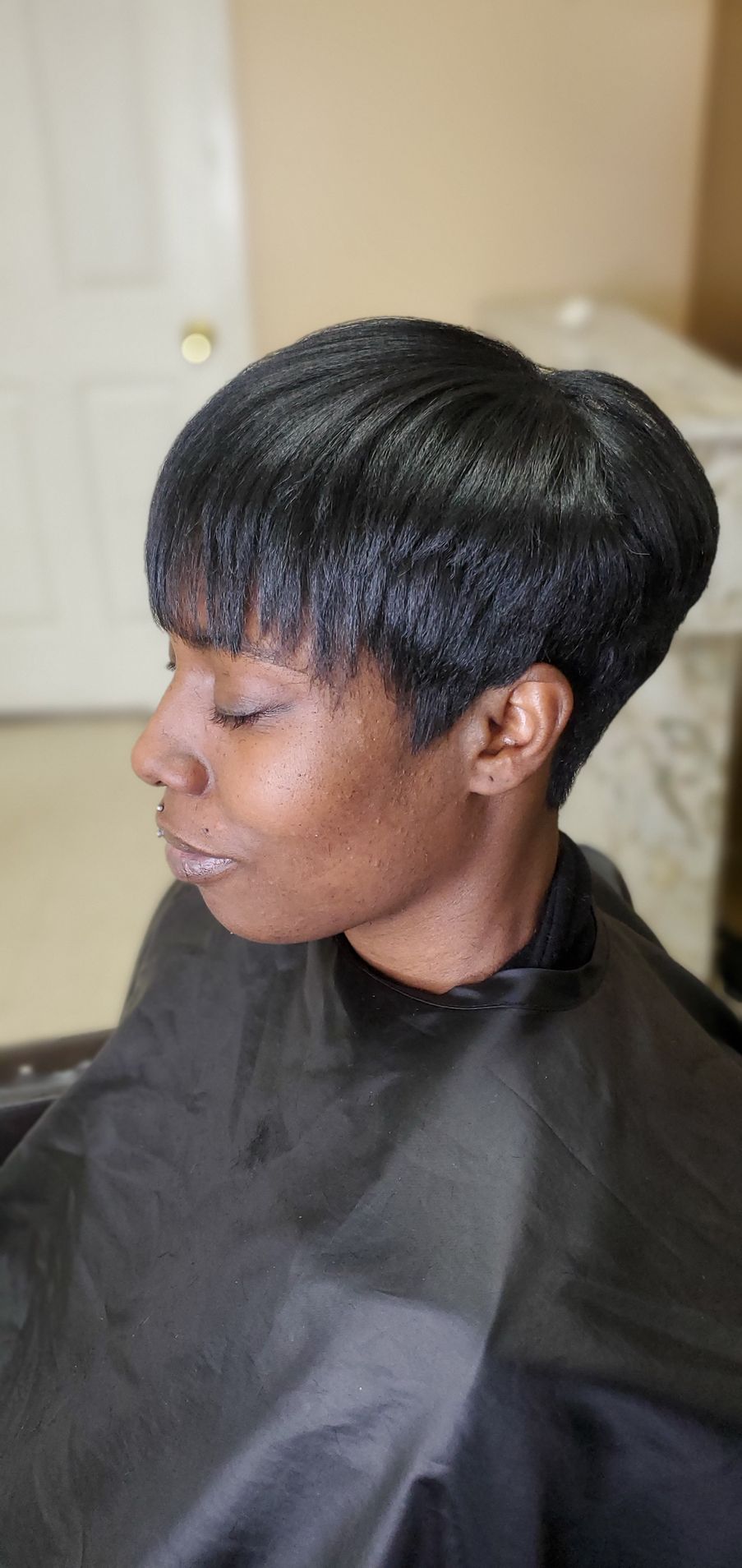 Side profile of a person with a short black haircut, wearing a black salon cape indoors.