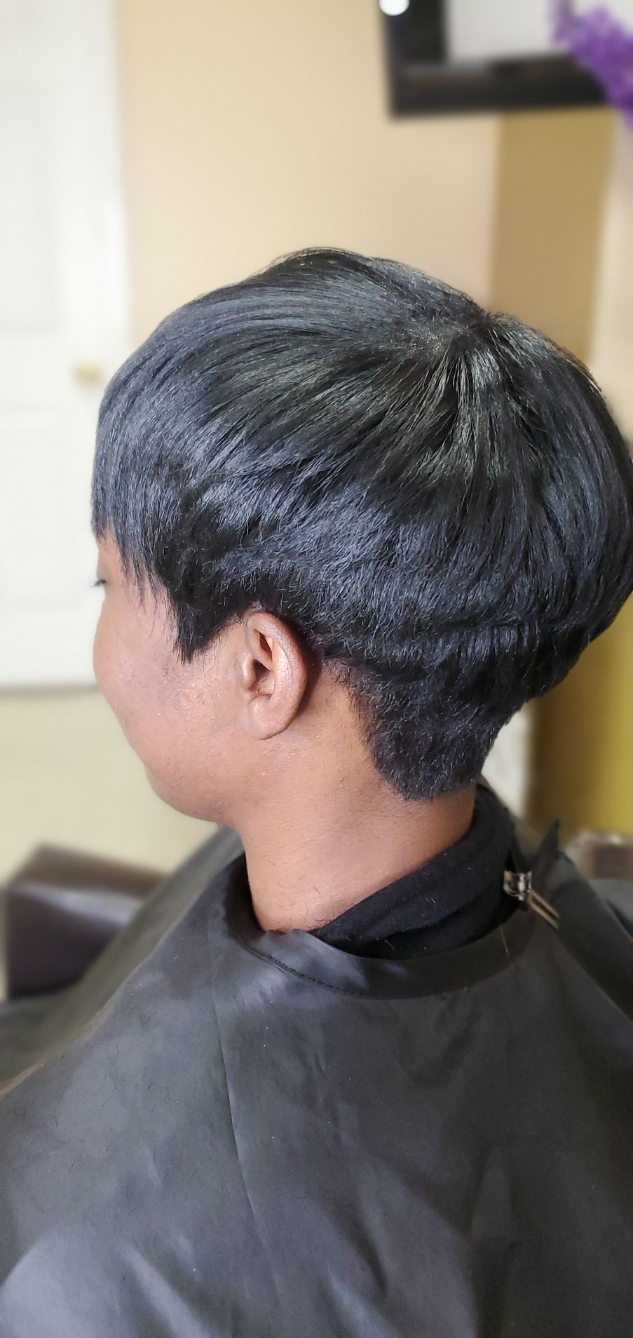 Side-view haircut with short, textured black hair, shown in a salon chair with a black cape.