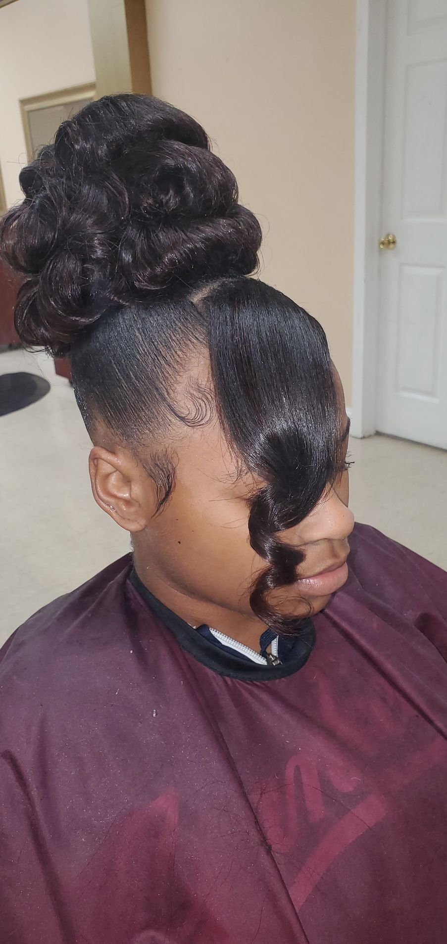Profile view of a person with styled updo and side curl in a salon chair, wearing a maroon cape