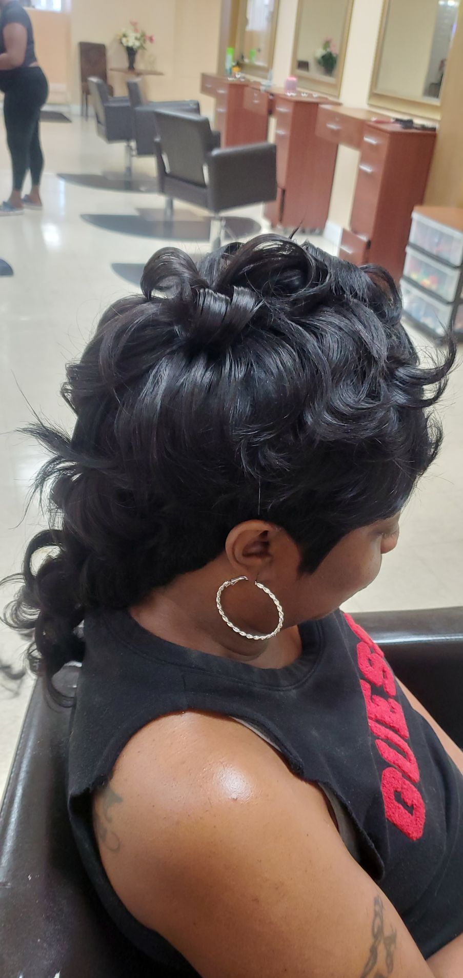 Woman with black curly updo and hoop earrings seated in a salon chair, seen in profile.