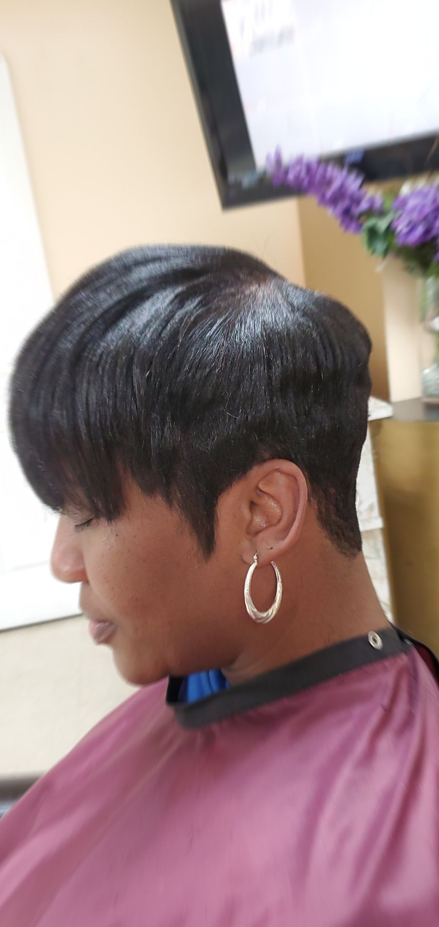 Profile view of a person with short black hair wearing a pink cape and silver hoop earring in a salon