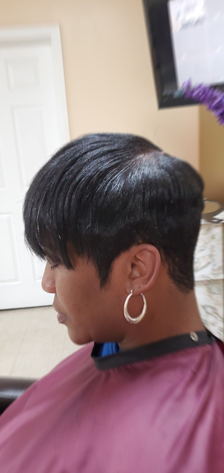 Person with short black hair and large hoop earrings, seated in profile in a salon chair wearing a maroon cape.