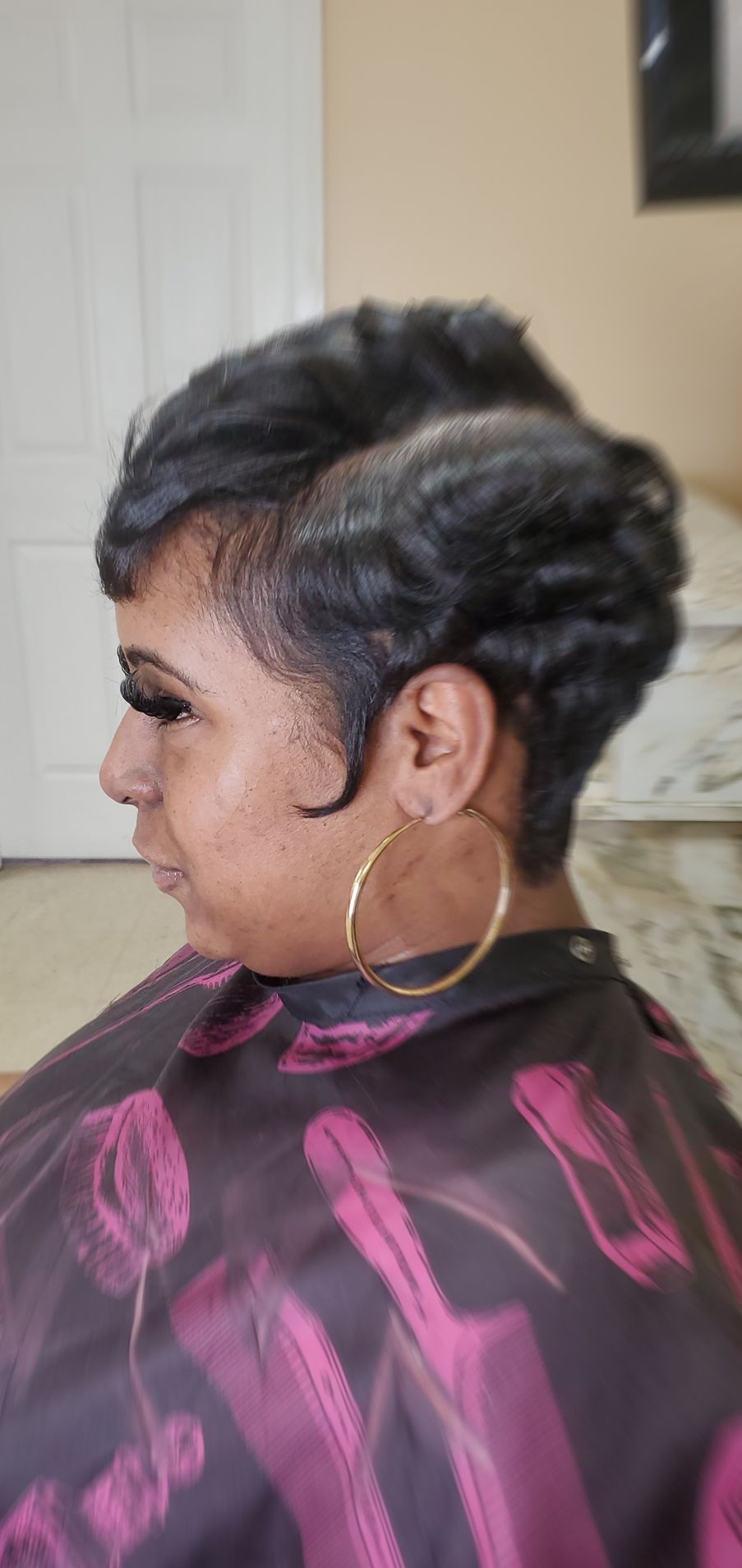 Side profile of a person with short dark hair and a hoop earring, wearing a patterned cape in a salon chair