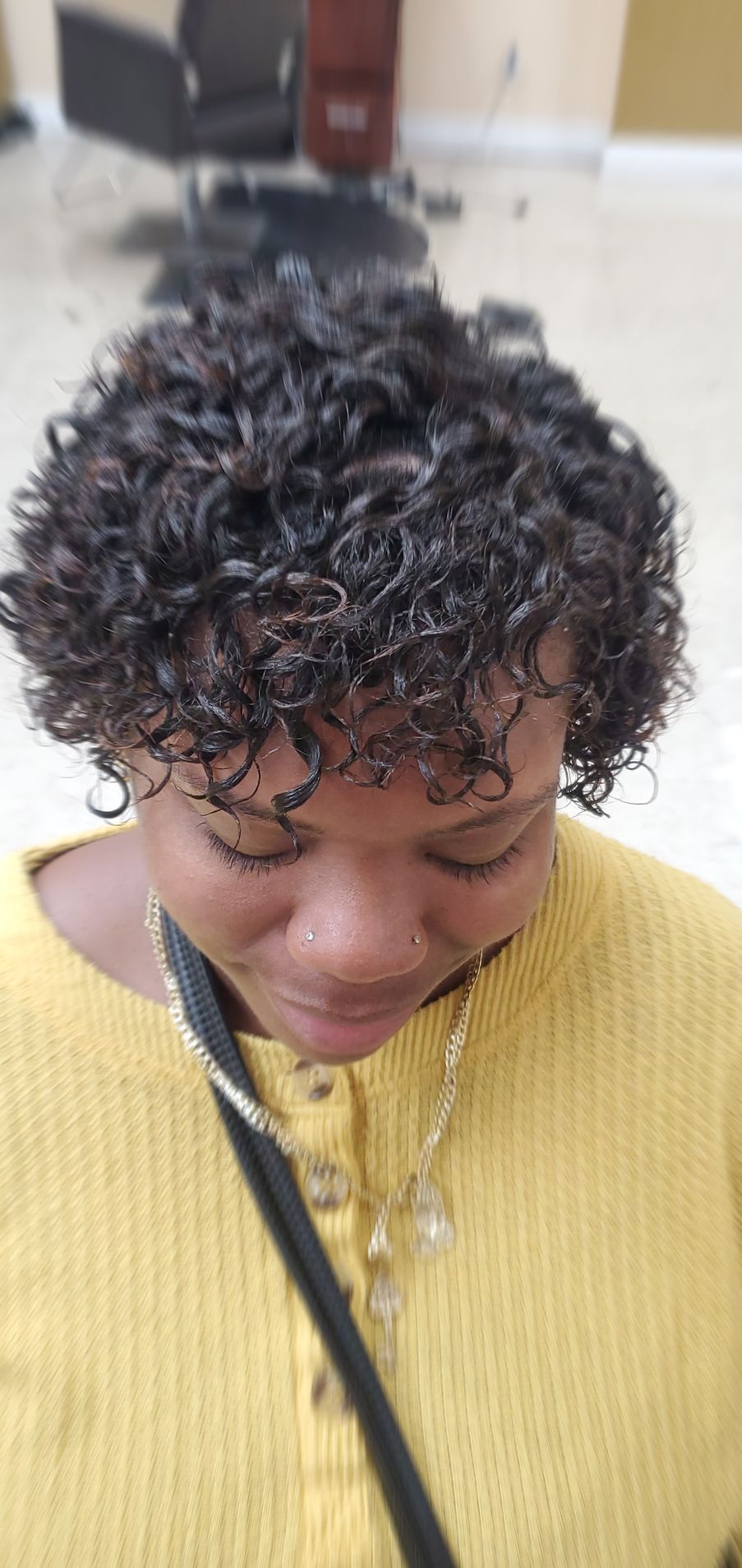 Person looking down in a yellow sweater, with short curly hair and a black shoulder strap indoors