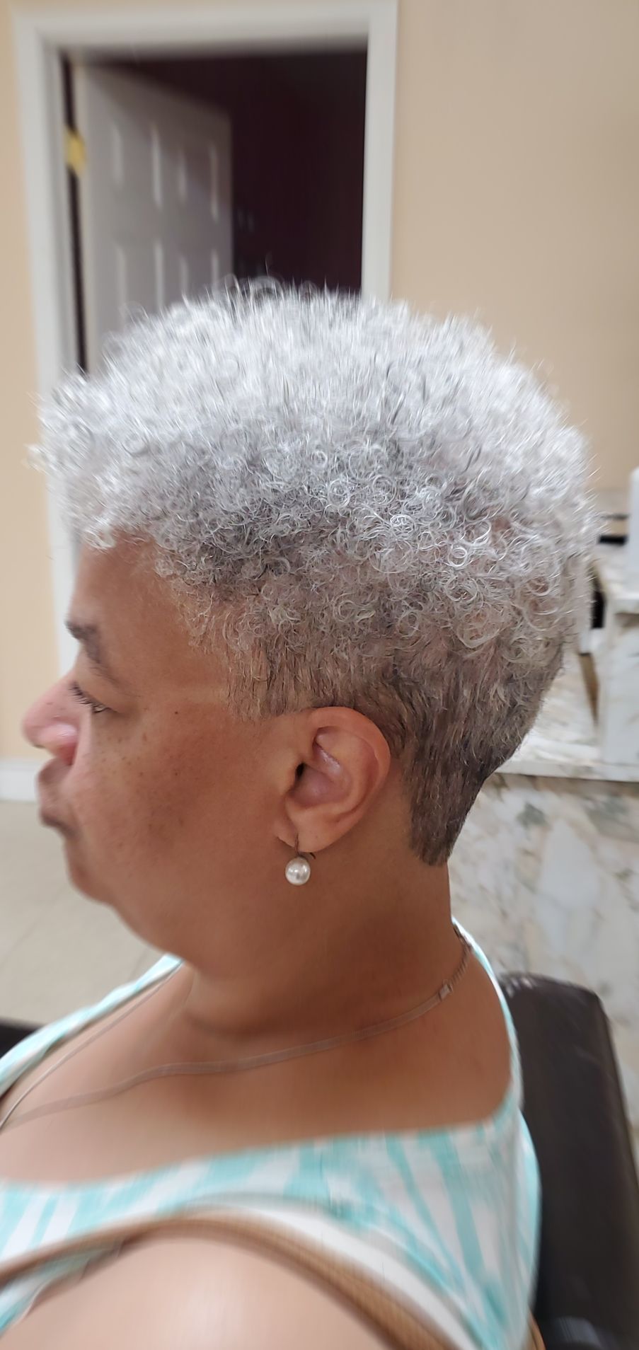 Profile view of a person with short white curly hair, wearing a turquoise top and pearl earrings indoors