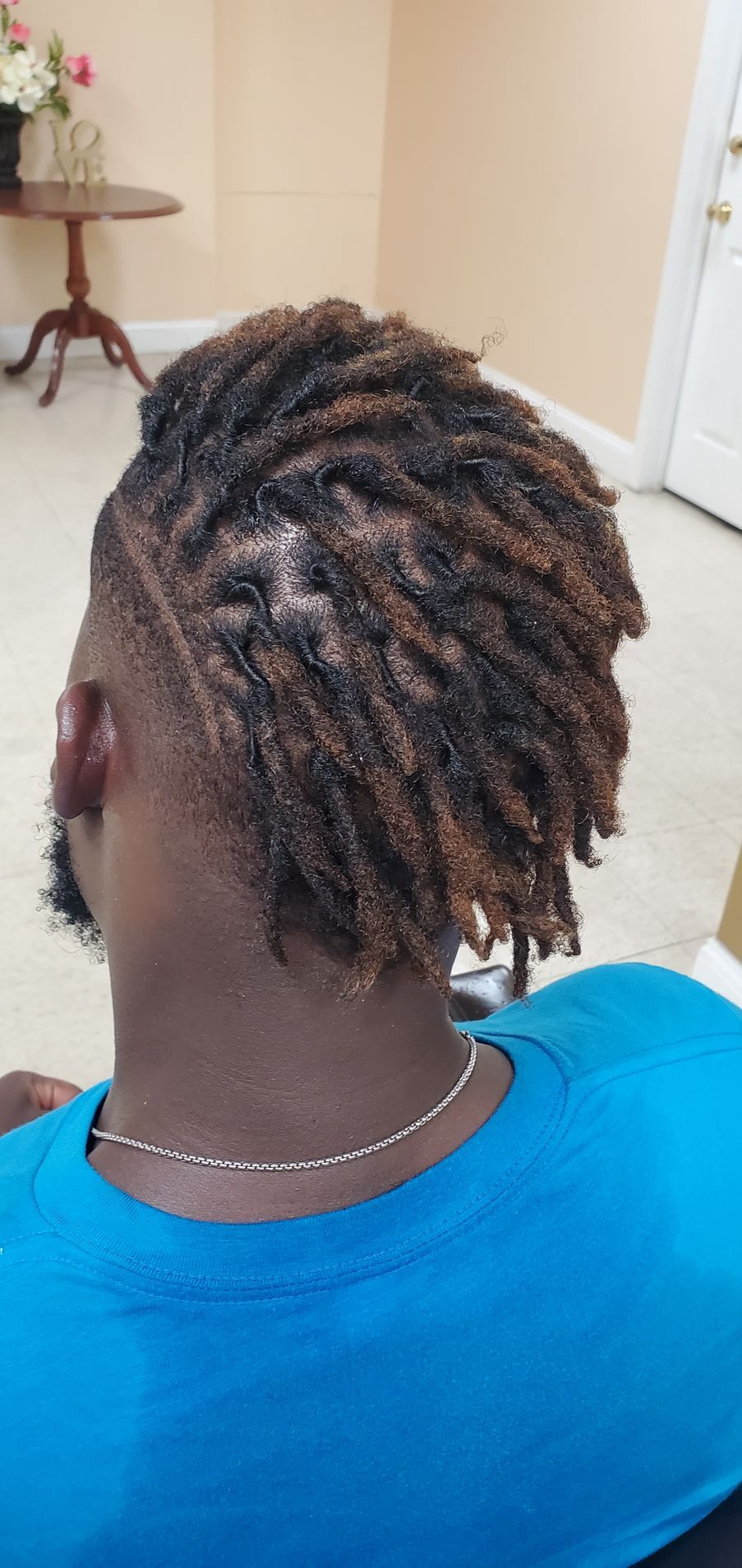 Back of a person’s head with short twisted braids, wearing a blue shirt indoors.