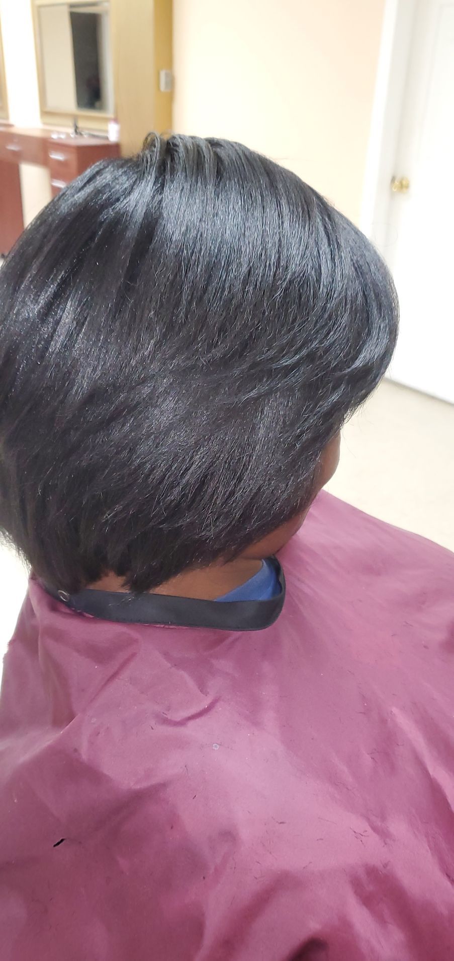 Person with short black hair wearing a purple salon cape in a bright room