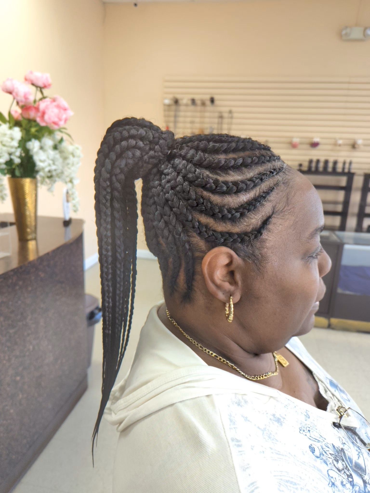Side view of a woman with long braided ponytail in a salon, wearing white and gold jewelry.
