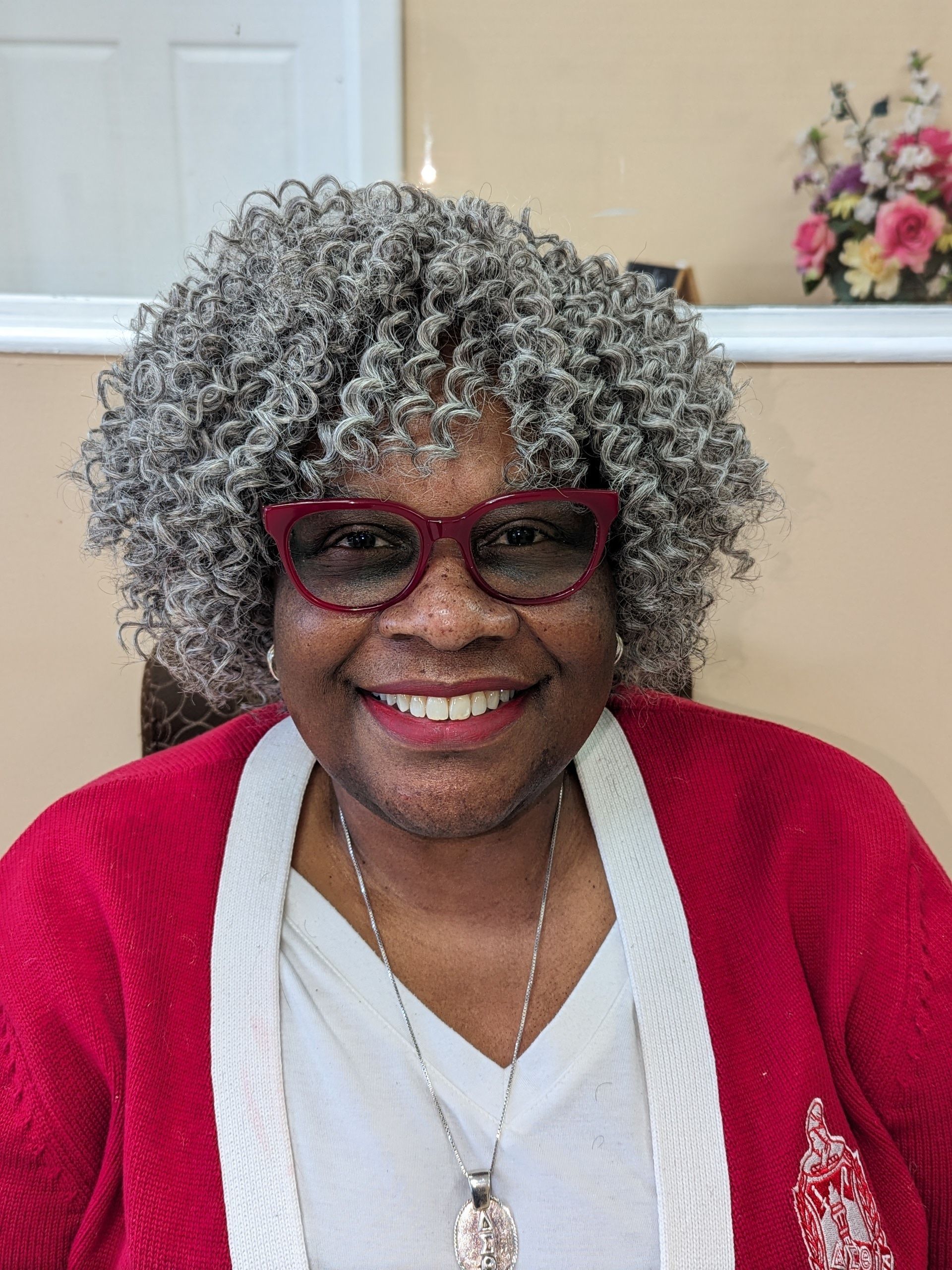Smiling woman in red glasses and cardigan, wearing a gray curly wig, seated indoors near flowers.