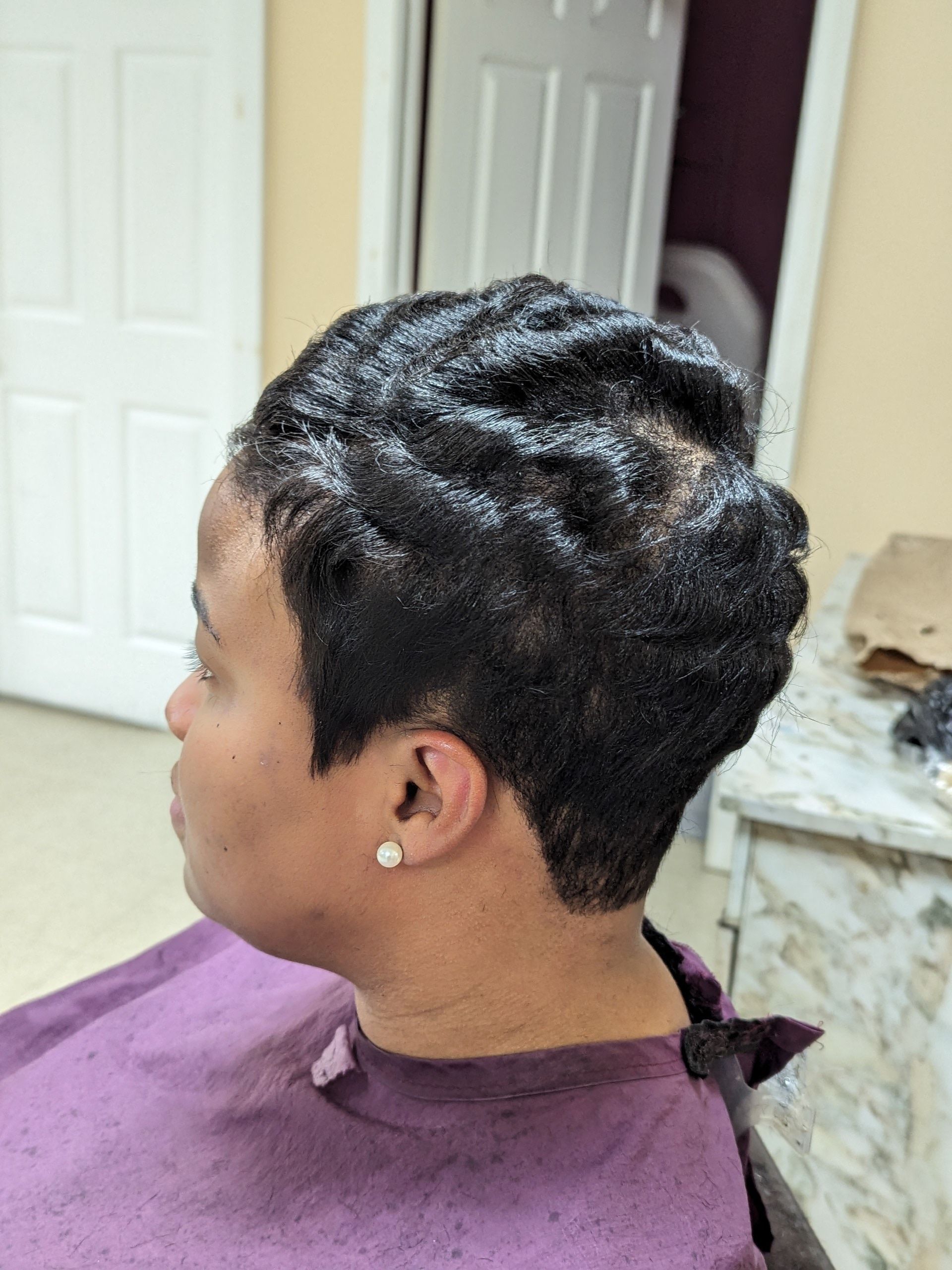 Side-profile of a person with short curly hair in a purple salon cape, seated indoors.