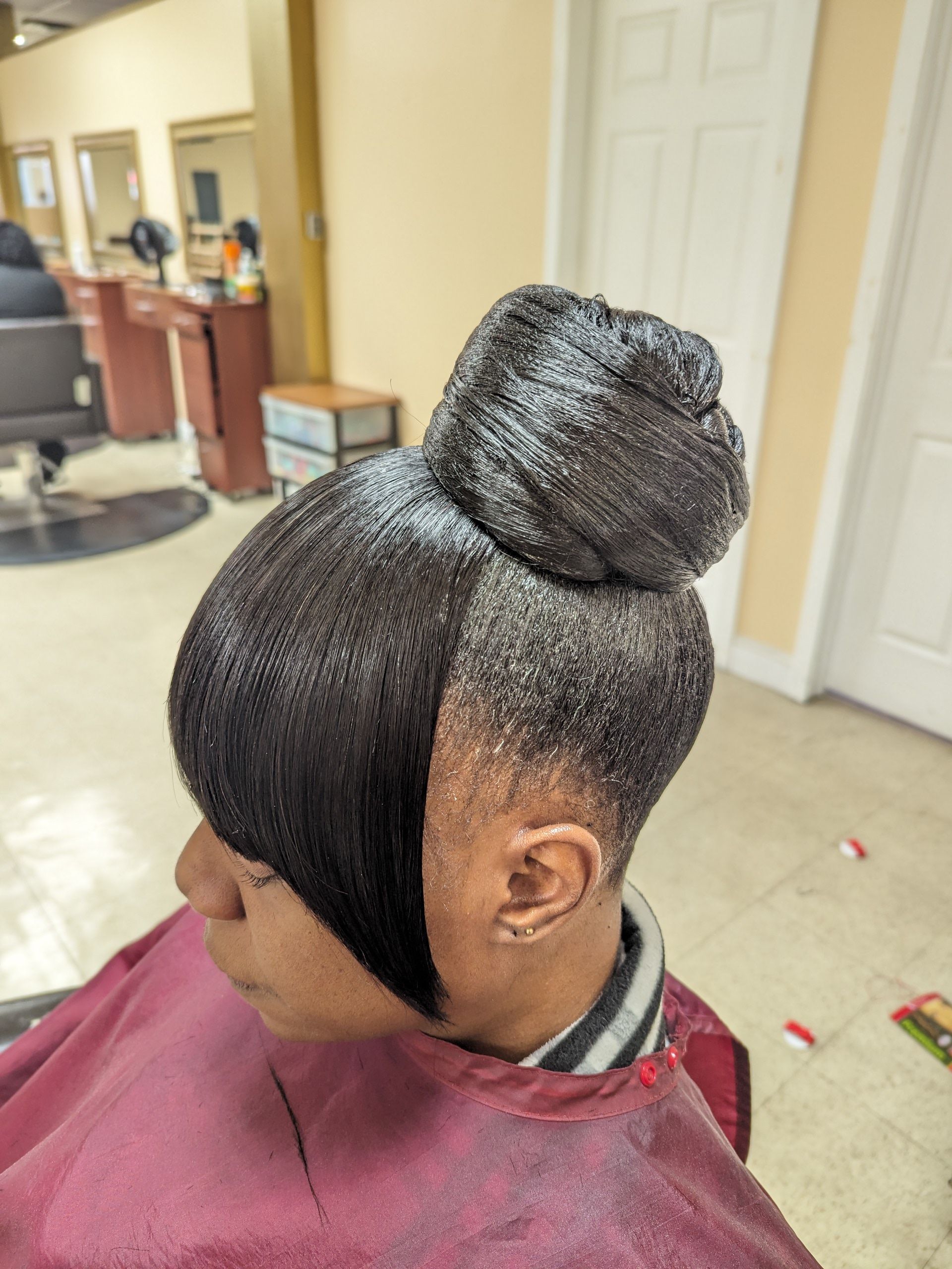 Side view of a person with a sleek braided bun, seated in a salon chair with a maroon cape.