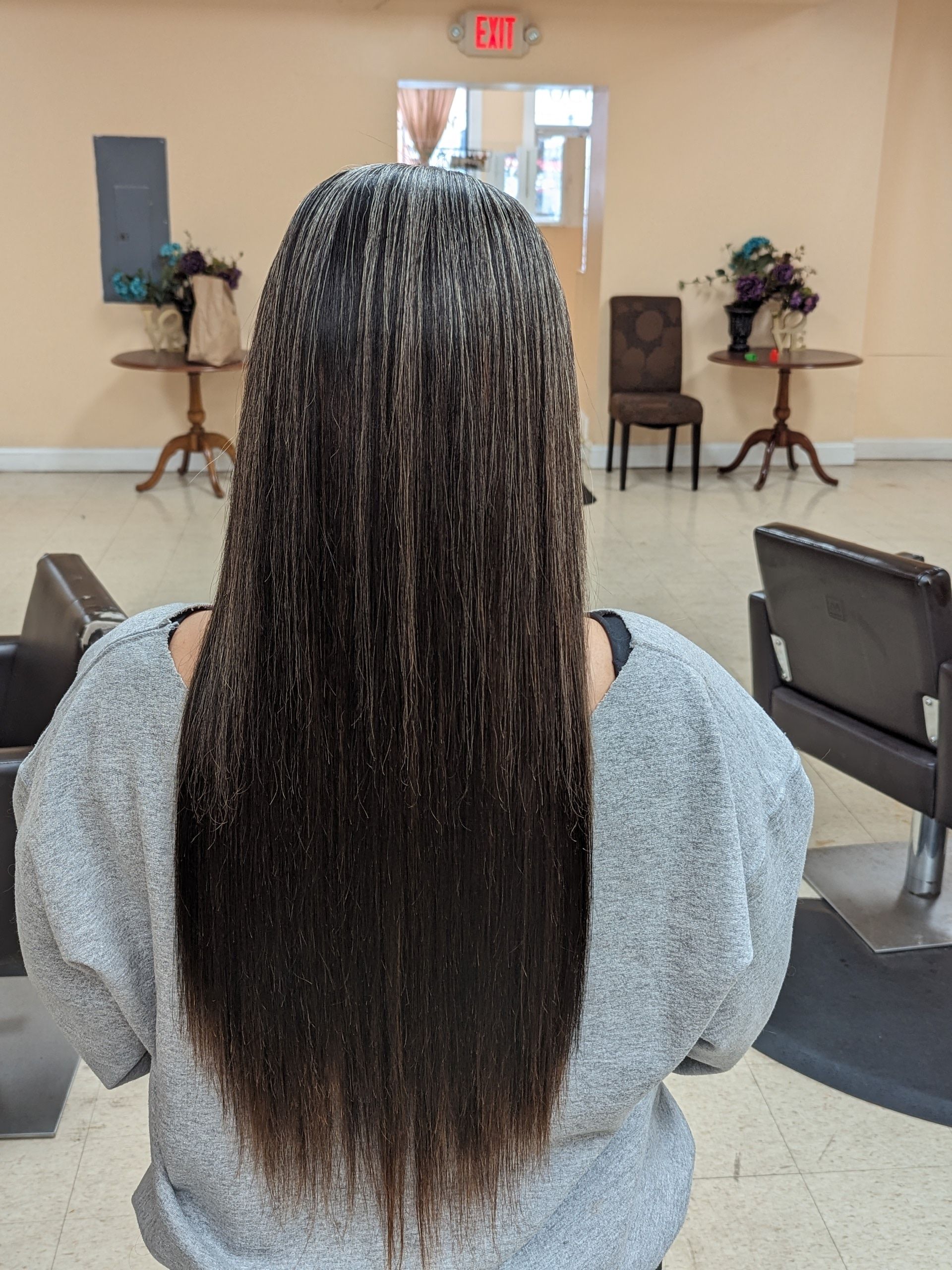 Person with long braided hair seated in a room, viewed from behind, with chairs and flowers in the background