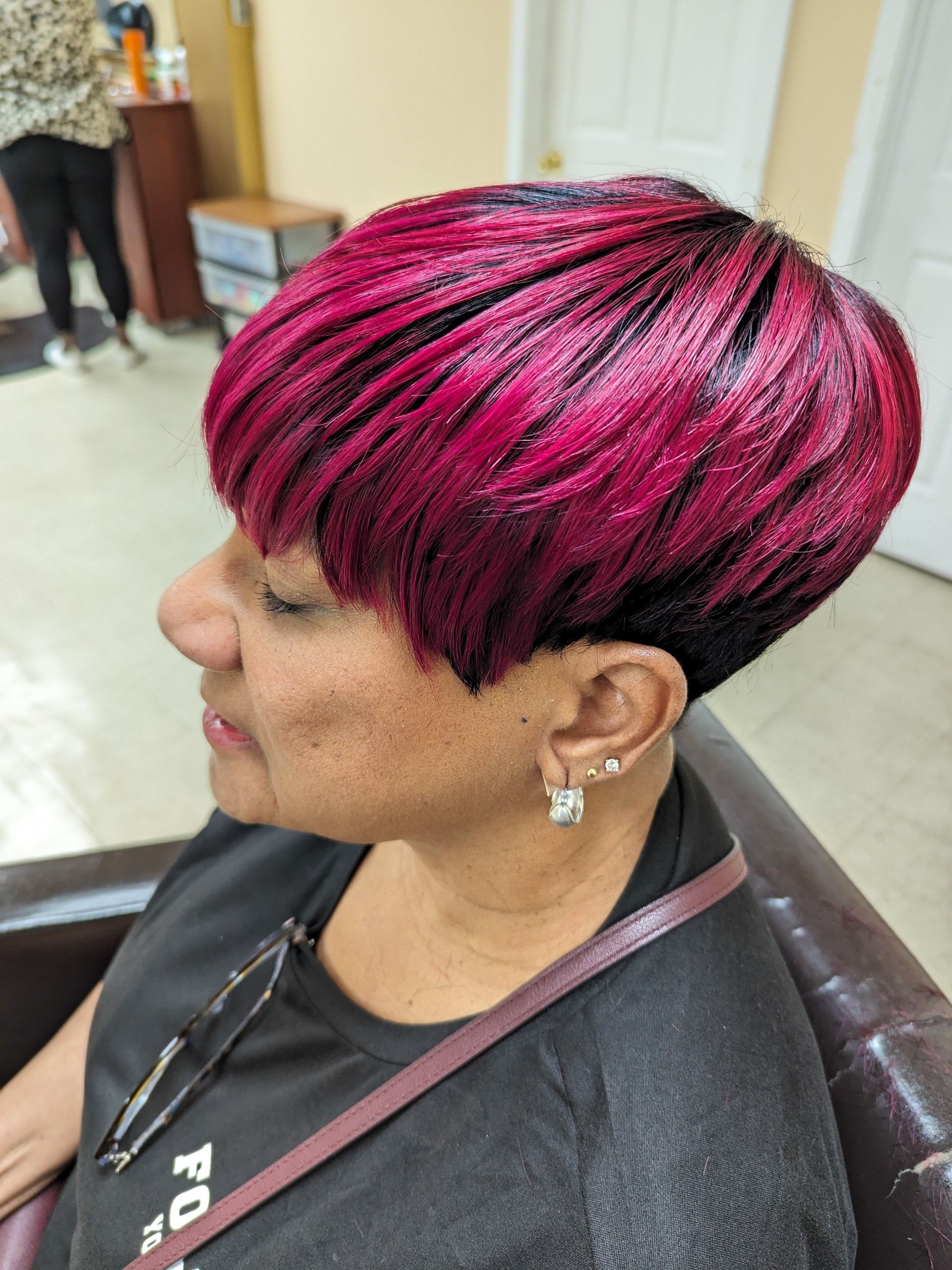 Side profile of a person with bright pink-and-black hair, wearing earrings and a black shirt in a salon chair.