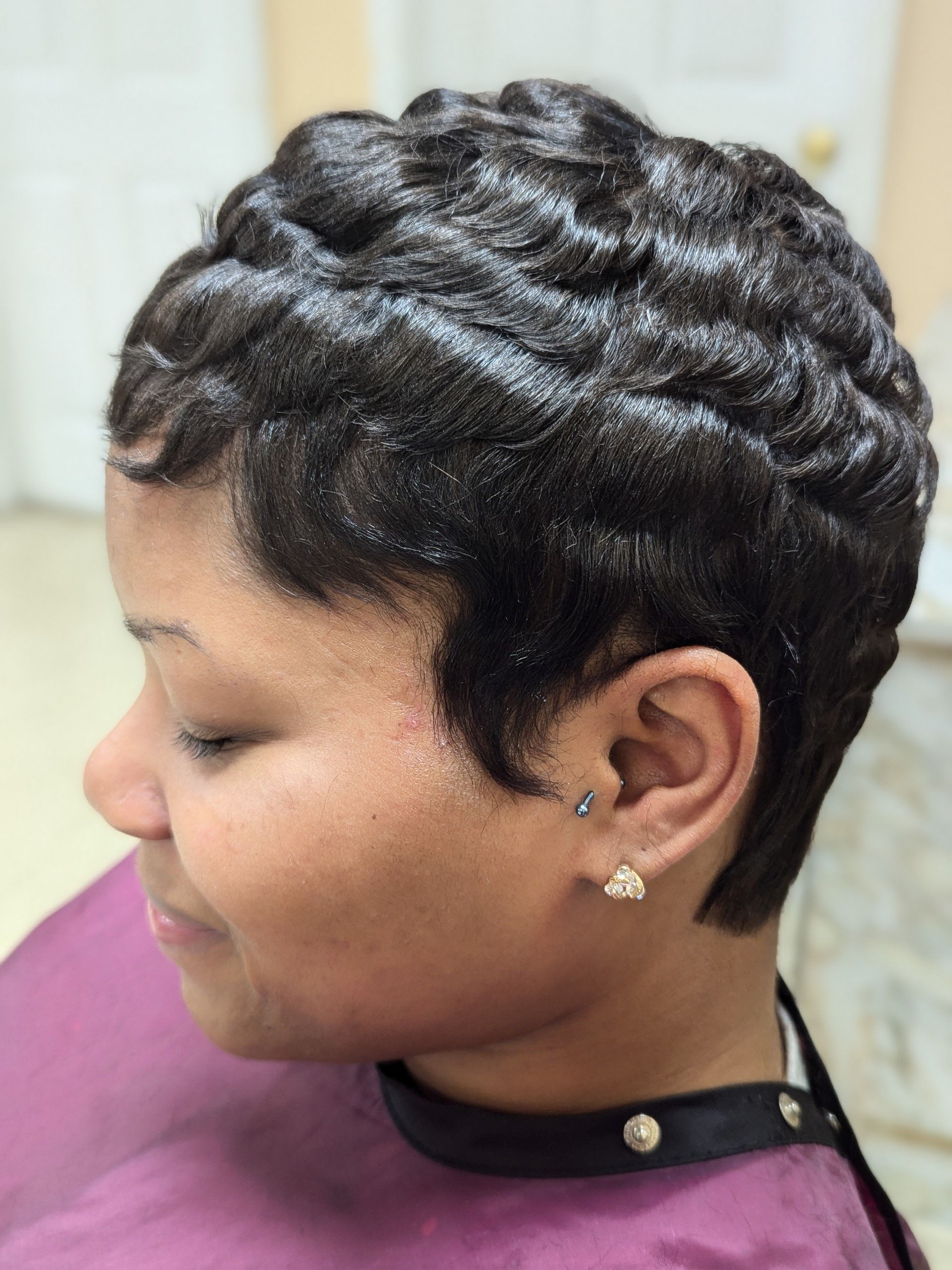 Side profile of a person with short braided hair, wearing a purple top and earrings, smiling gently.