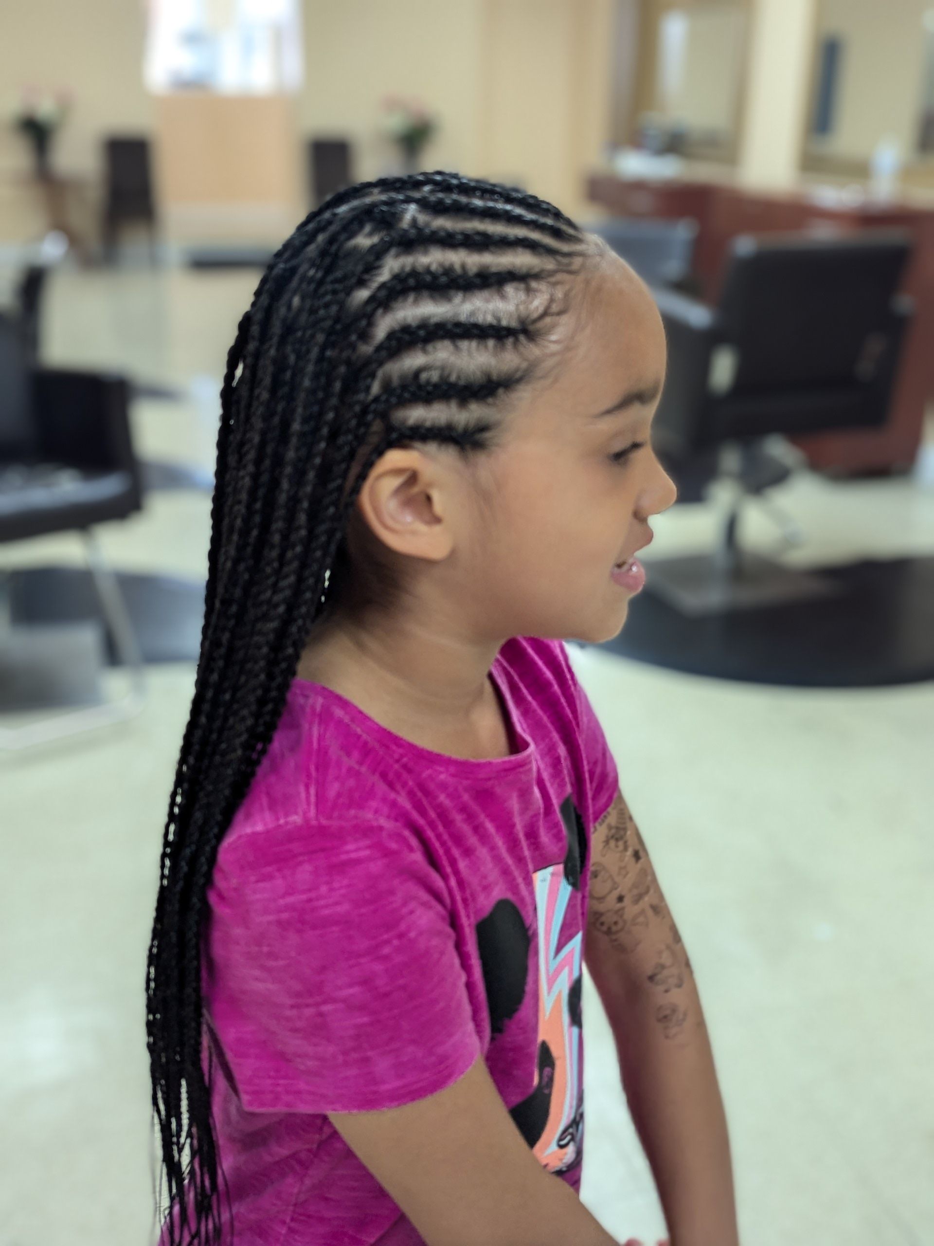 Young child with braided hair in a pink shirt, seated in a salon chair, side profile.