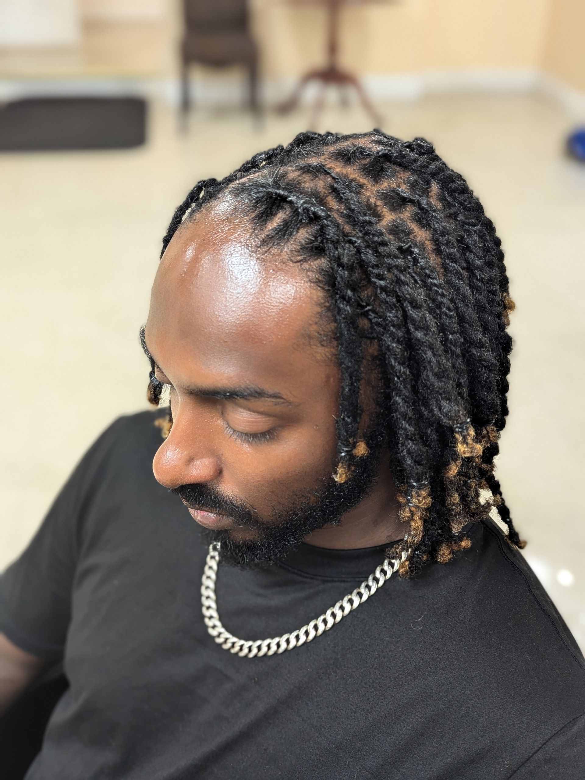 Man with braided hair looking down, wearing a black shirt and silver chain necklace indoors