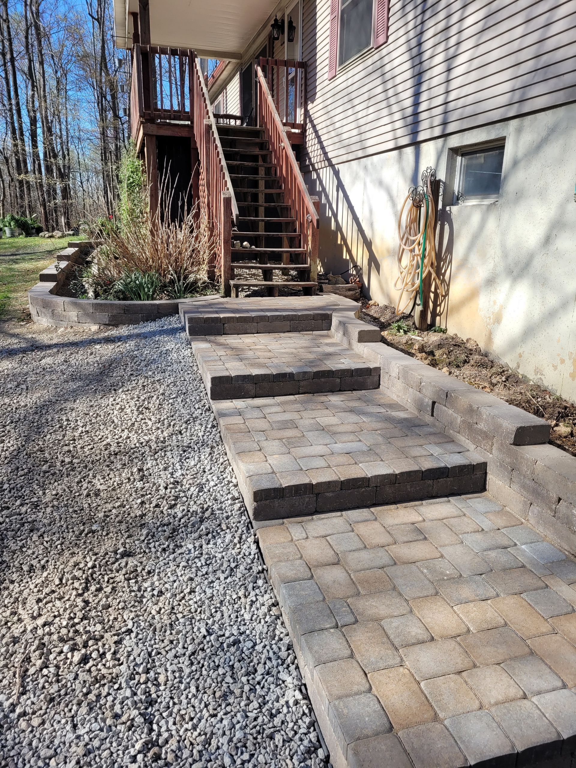 A set of stone steps leading to a wooden deck, flanked by a low retaining wall and a gravel path beside a building.