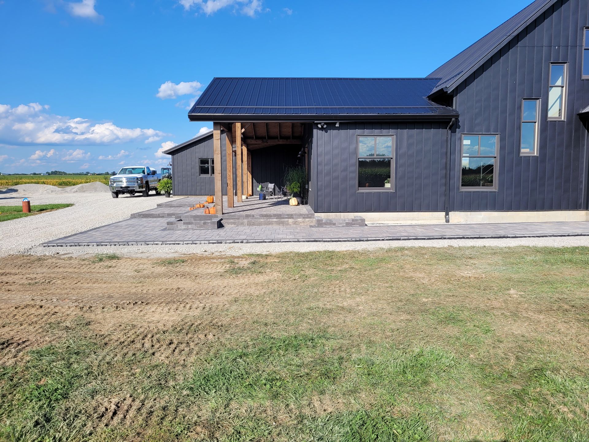 A modern black farmhouse with a covered patio, gravel driveway, and a truck parked under a blue sky.