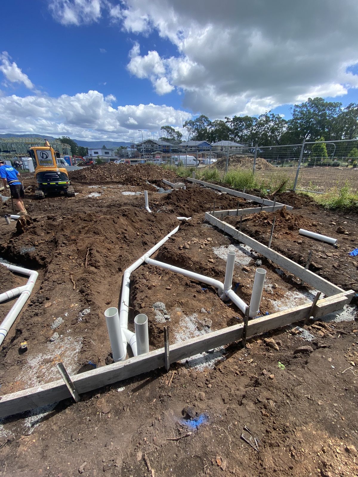 Construction site showing a trench foundation with exposed white plumbing pipes under a bright, cloudy blue sky.