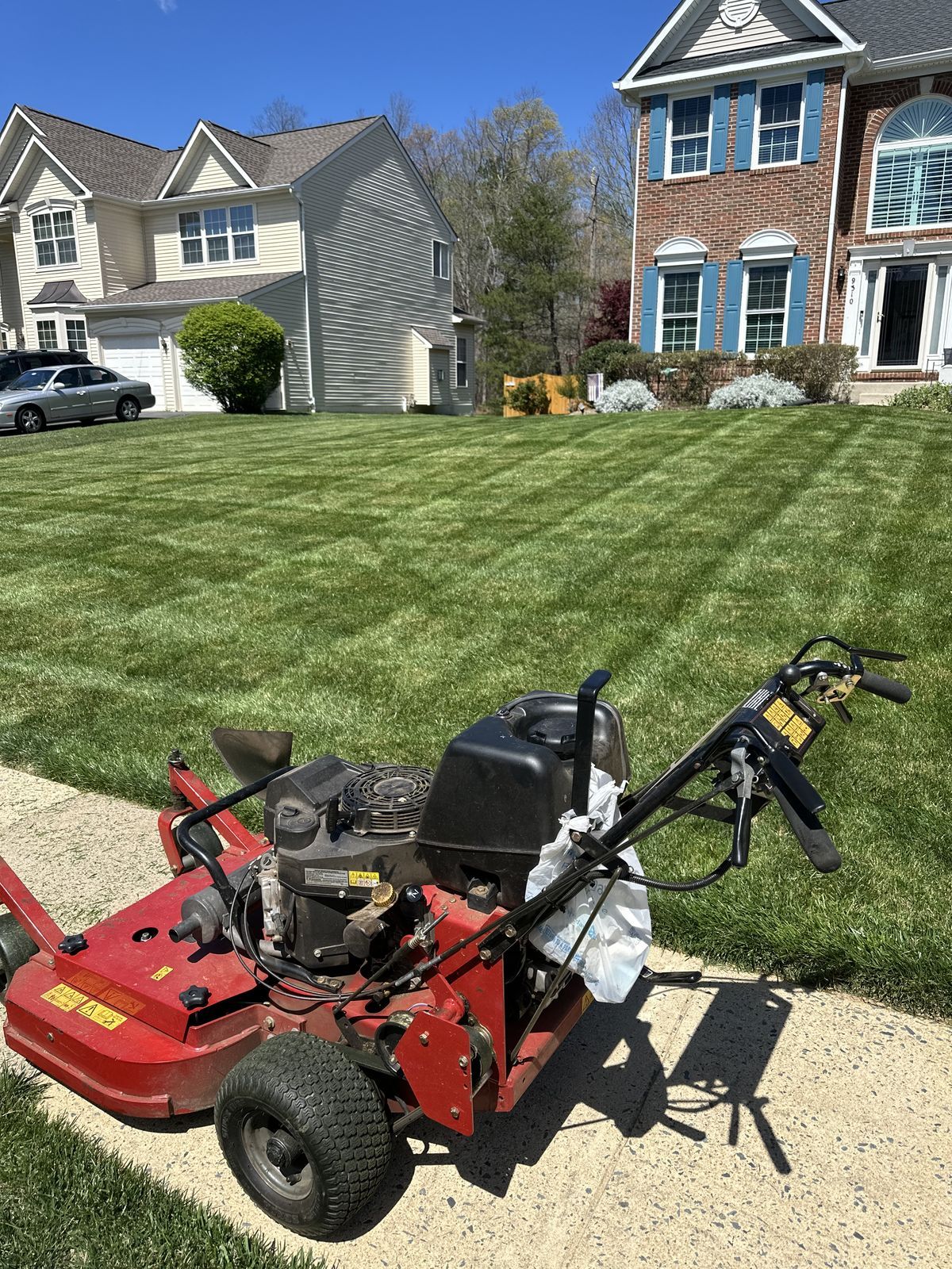 A red commercial walk-behind lawn mower parked on a sidewalk in front of houses with freshly mown, striped grass.