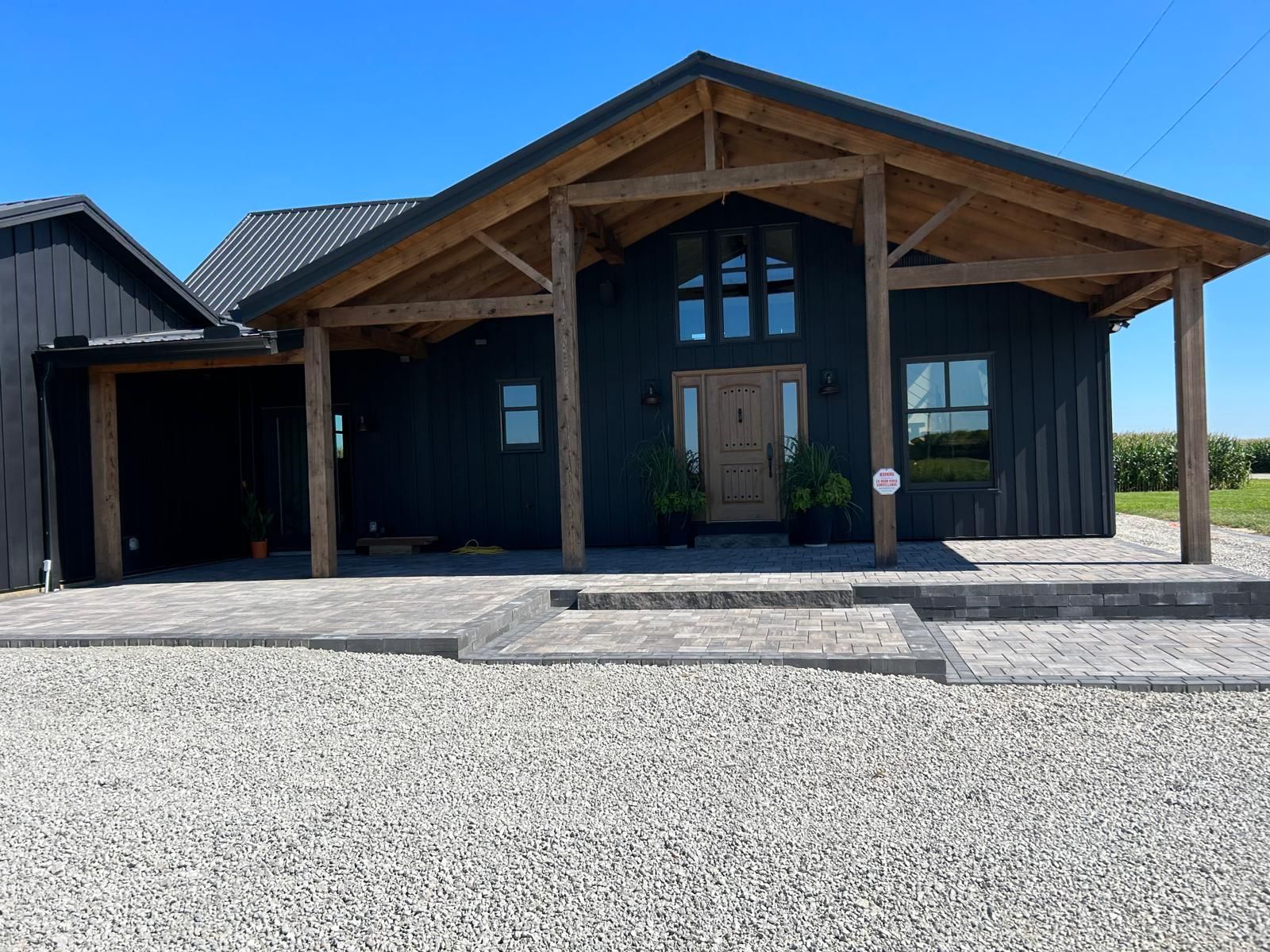 A modern black house with natural wood framing and a covered porch entrance, featuring stone patio flooring and gravel.