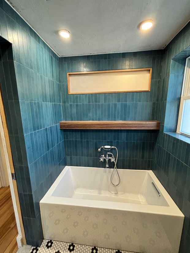 Small bathroom with teal tile walls, a white soaking tub, and a wooden shelf under a window.