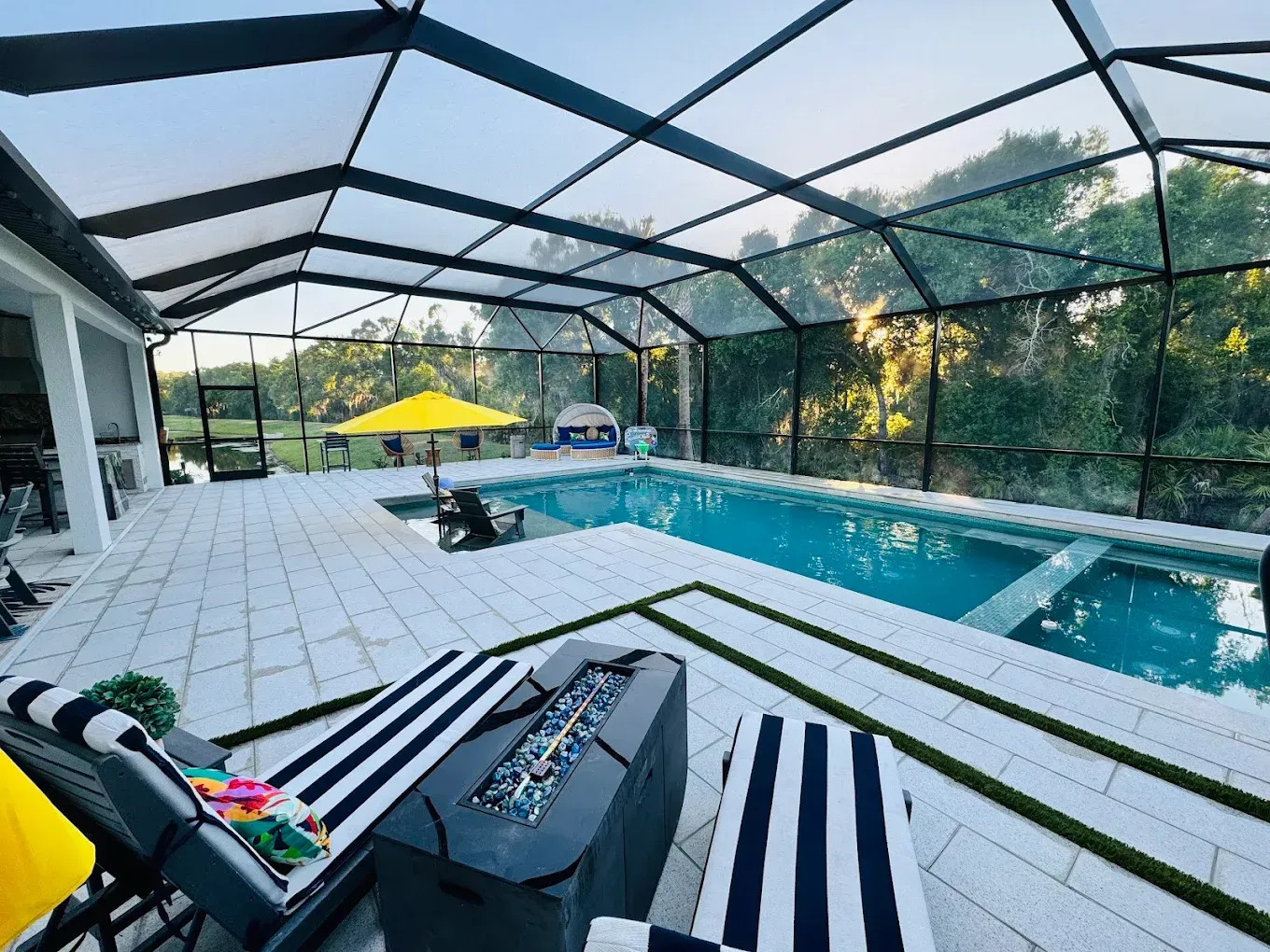 Pool and patio with a black-screened enclosure. Two striped lounge chairs, a fire pit, and a yellow umbrella are visible.