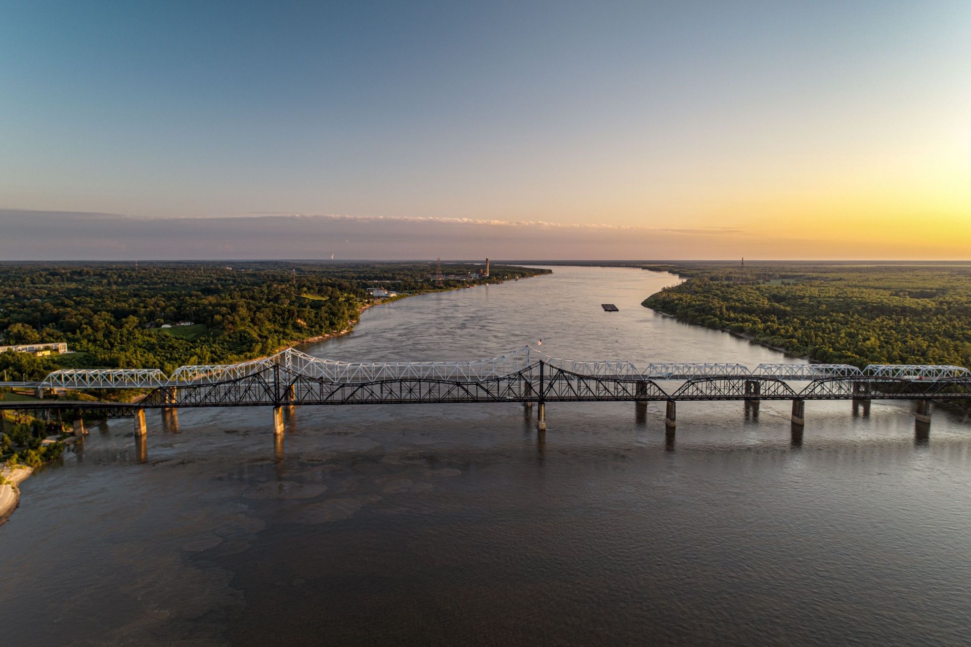 A bridge spans a wide river at sunset, with forested banks and a glowing sky.