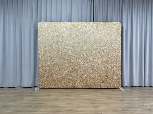 A gold glitter backdrop is sitting on a wooden floor in front of a curtain.