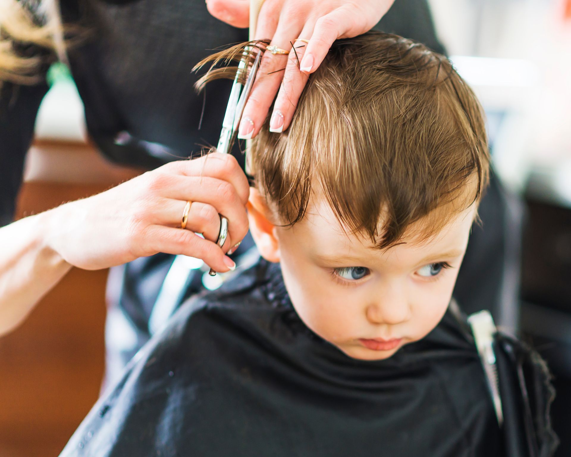 Child getting a haircut, held by stylist. Boy with blue eyes, black cape, scissors cutting hair.