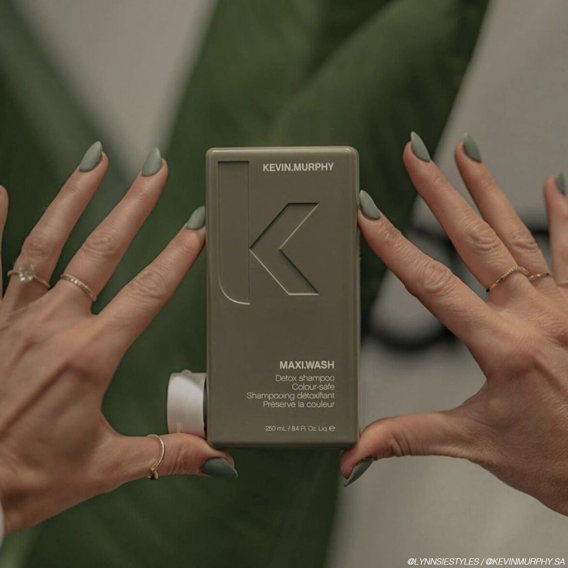 Hands with green nails holding a green hair product container against a green backdrop.