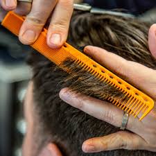 Hands of a barber combing and cutting a person's dark hair with orange comb and scissors.