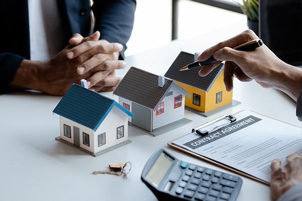 A person pointing a pen at three model houses on a table with a contract and calculator, suggesting a real estate deal.