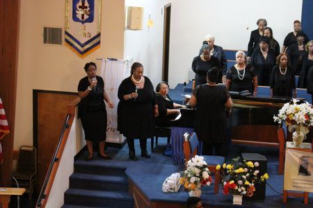 Choir singing in a church sanctuary, with a conductor at the front and floral arrangements on stage