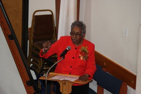 Person in a red jacket speaking at a podium beside a microphone in a church or hall