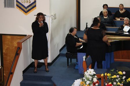 Woman speaking at a church podium while others sit and sing at a piano during a service