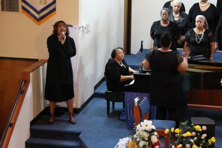 Choir singing in a church, with a soloist standing by a piano and flowers in the foreground