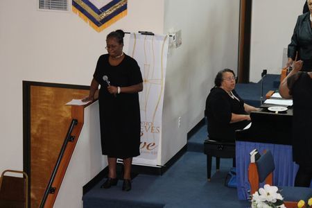 Woman speaking at a podium in a church, with another woman seated at a piano nearby.