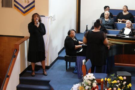 Woman singing at the front of a church during a service, with musicians and choir behind her.
