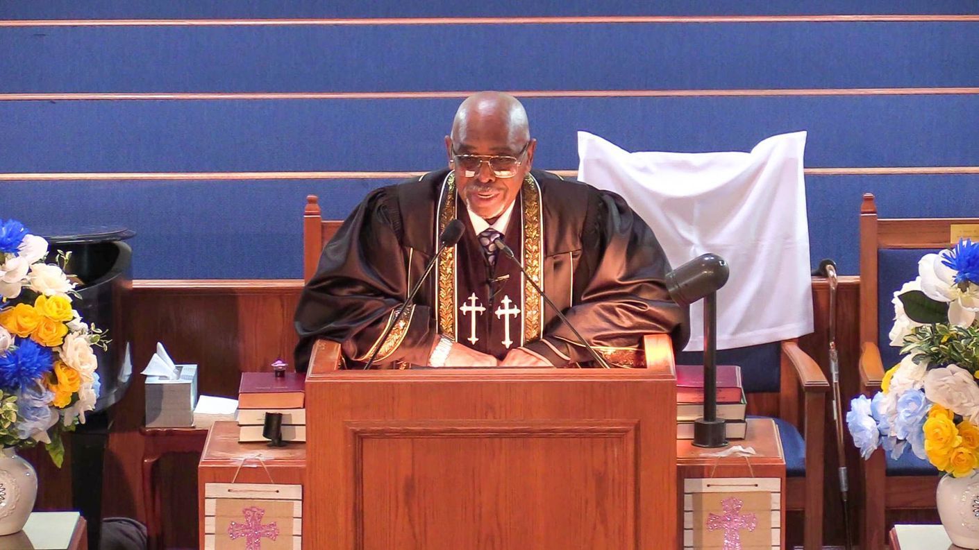A Pastor stands at a wooden pulpit in a church, reading under blue walls and floral arrangements