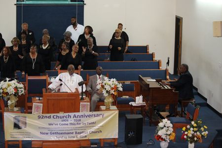 Church anniversary service with choir on blue pews and speaker at pulpit behind floral decorations.