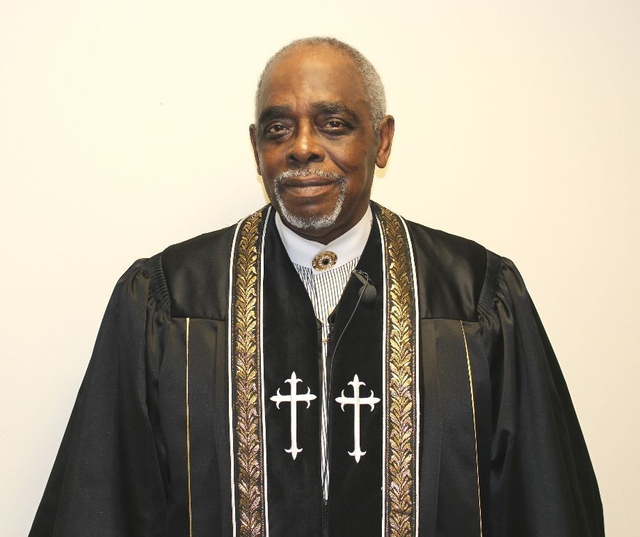 Pastor in black clerical robe with white crosses, standing against a plain light background