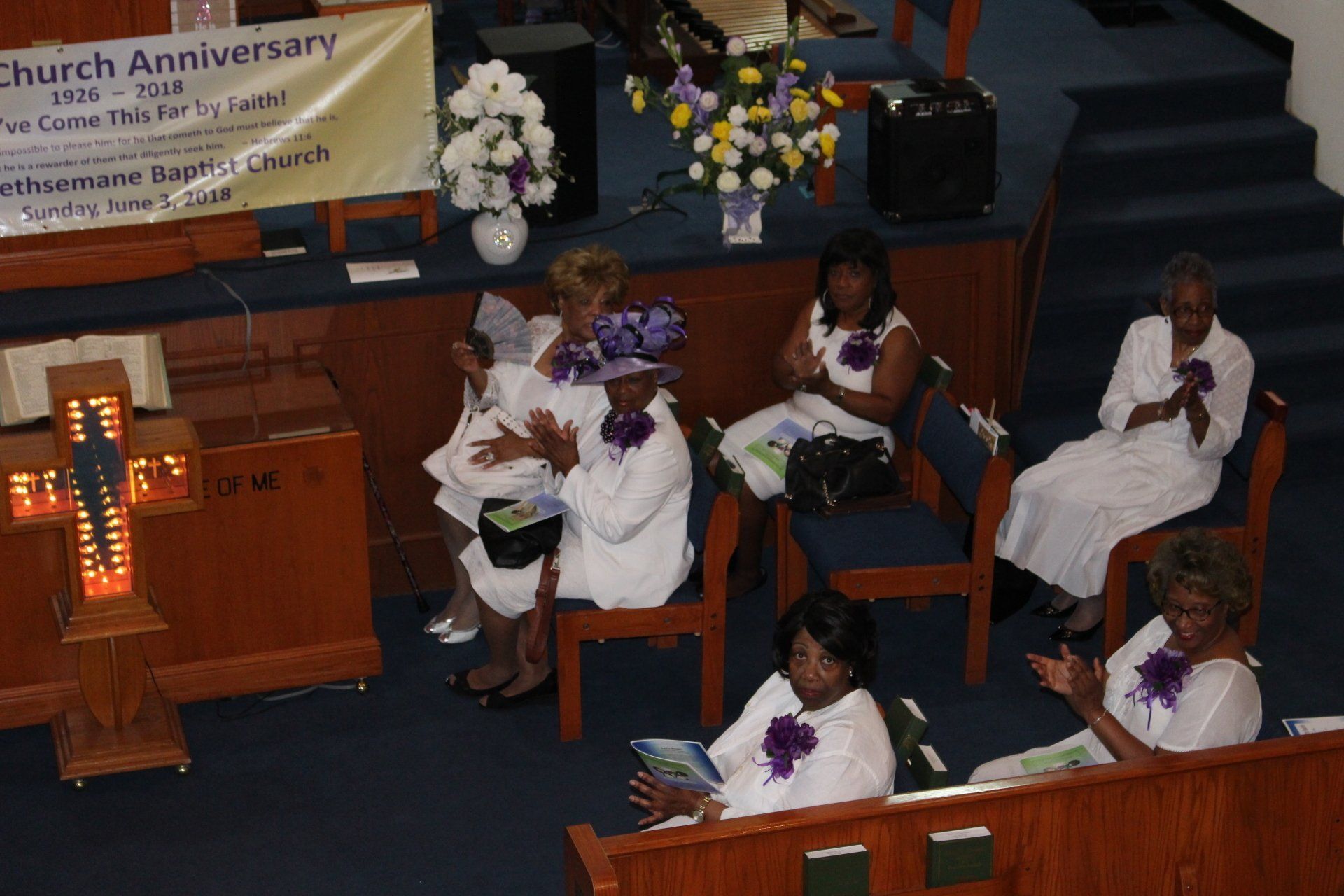 People in white robes seated in a church sanctuary during a service with flowers and a podium